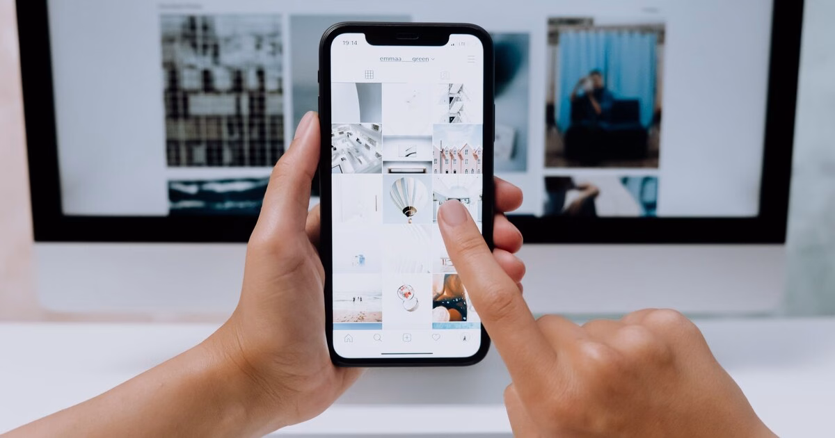 Hands holding a smartphone browsing images in front of a desktop monitor